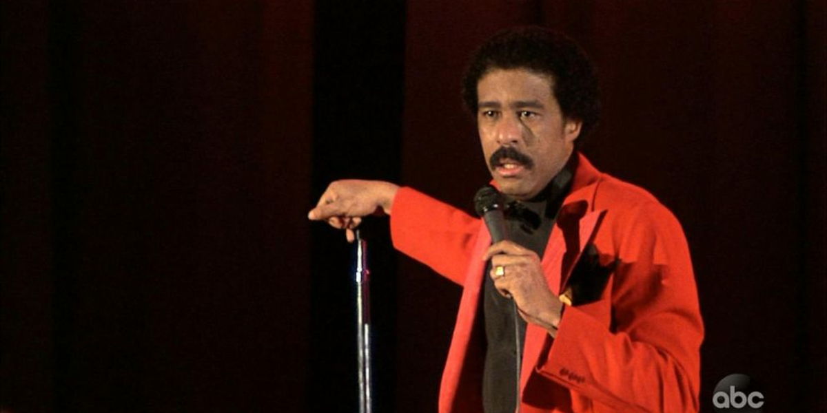 Richard Pryor in a red jacket standing at a microphone on a dark stage.