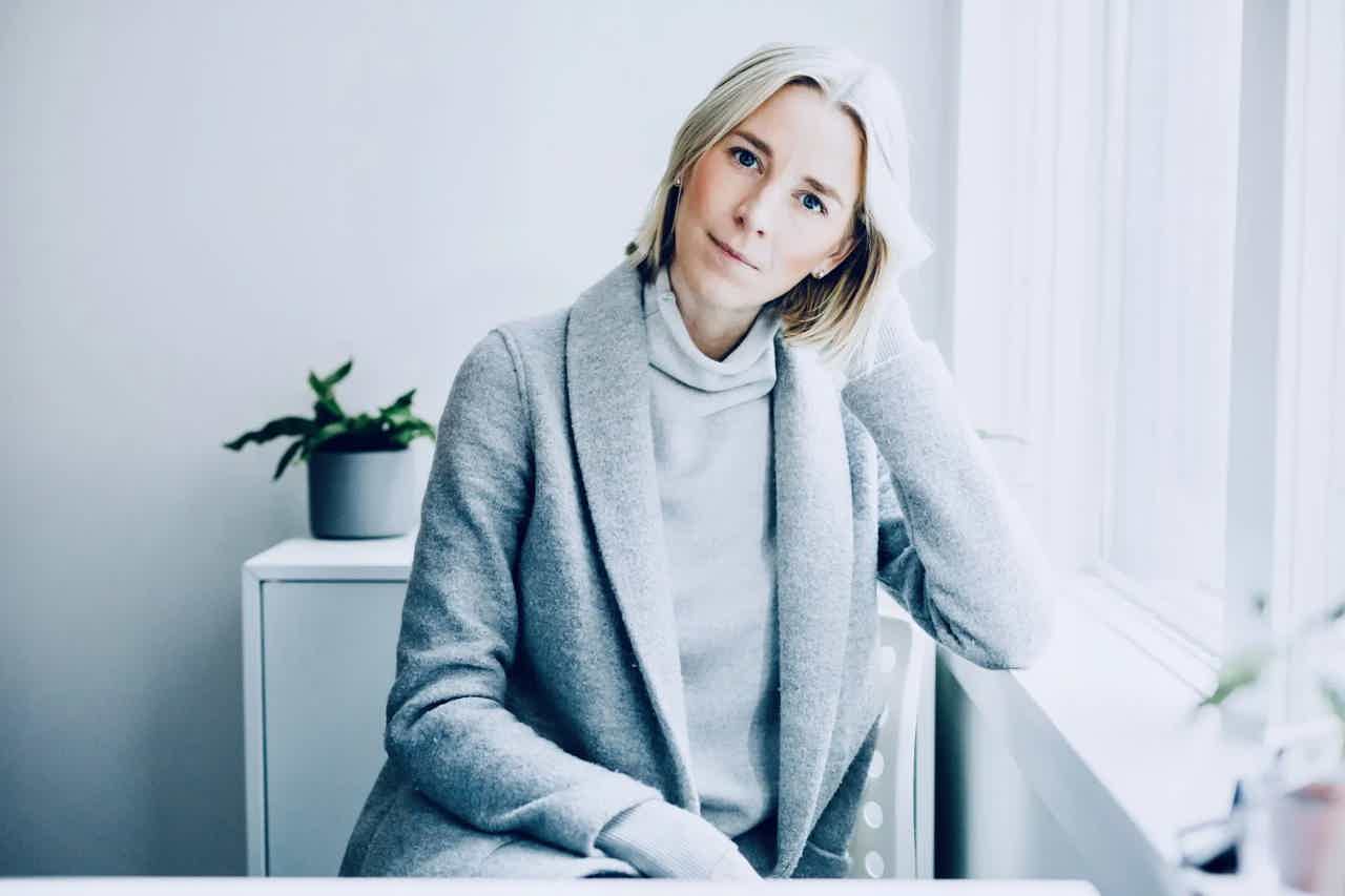 Woman with blond bob wearing gray coat and light turtleneck sits by window, head tilted, soft natural light.