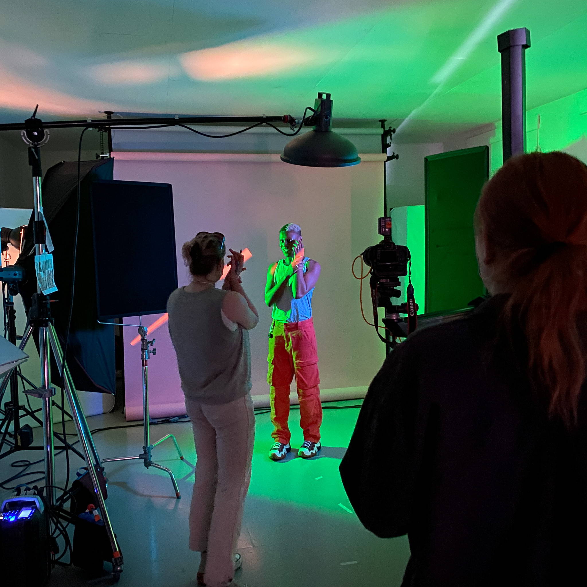 Model Kenya can be seen in the centre of the image wearing bright red cargo trousers and a light coloured tank-top. They are holding their lright arm up with their left to the side of their face. Camera lighting equipment stands to the left. In the foreground, two stylists are facing Kenya. 