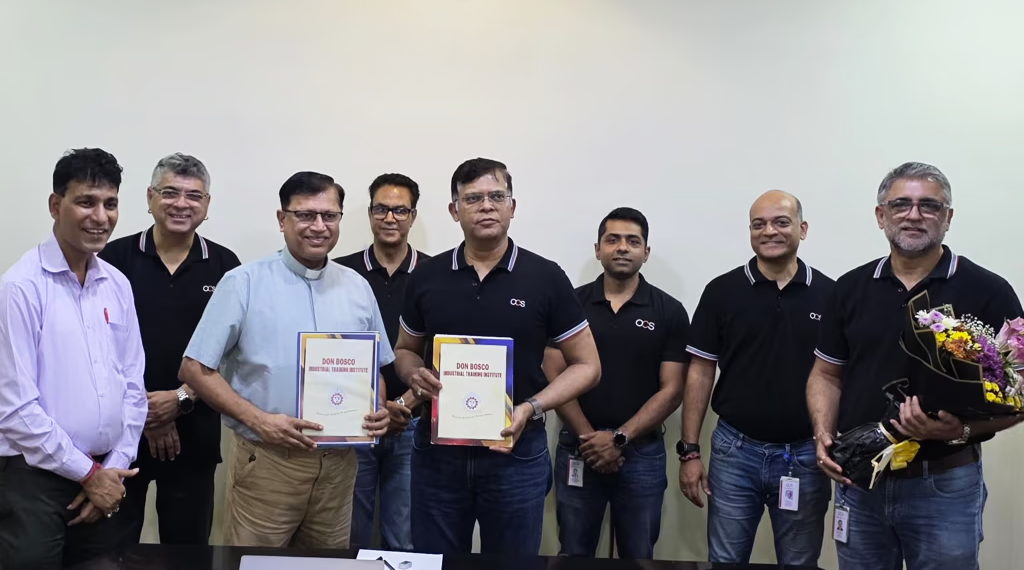 Eight men stand indoors, most wearing black polo shirts with a company logo. Two men in the center hold documents, while the man on the right holds a bouquet of flowers. All are smiling and facing the camera.
