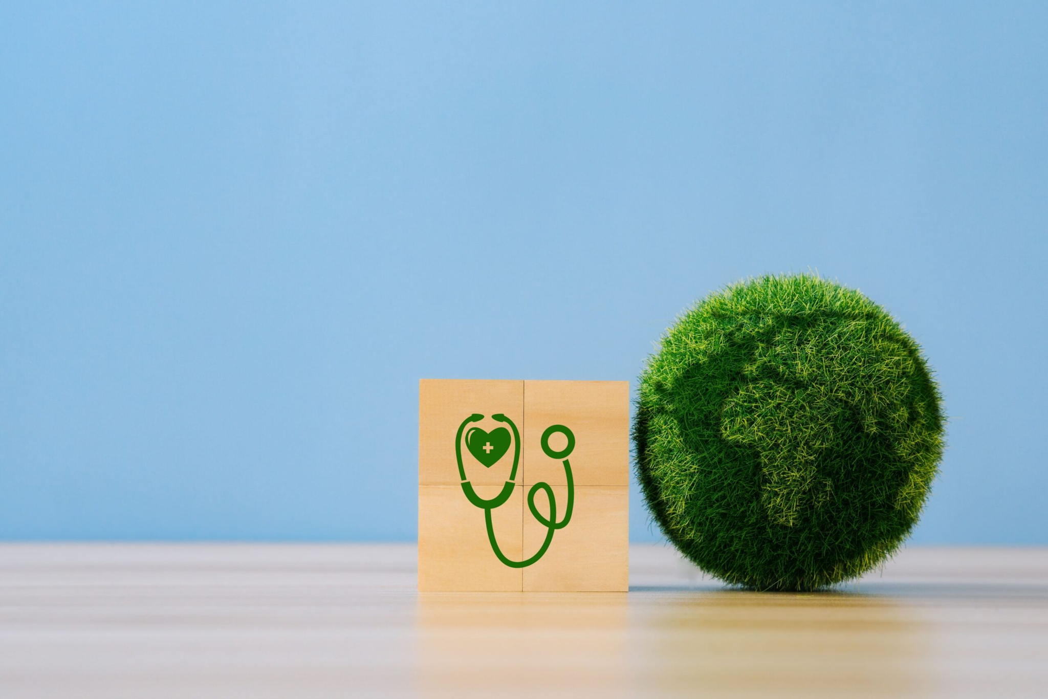 A green grassy globe sits next to four wooden blocks with a green stethoscope icon, symbolizing global health and environmental care, on a light wooden surface against a blue background.