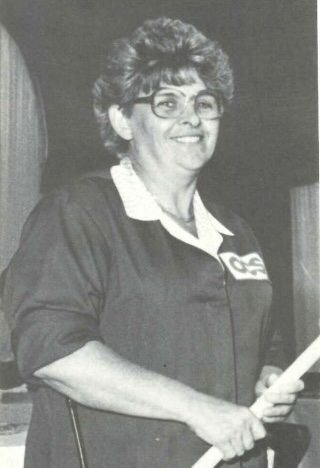 A person with short hair, wearing glasses and a dark collared shirt with a patch, holding a cue stick. The background is dark, possibly in an indoor setting. The person is smiling.