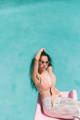 a woman with long black hair, pink sunglasses, and a pink crop top sits on the bottom of a pink slide above the pool below for poolside pride: a slim aarons-inspired pride campaign