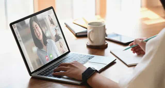 A person on a laptop screen in a video call with multiple participants, writing notes on a pad.