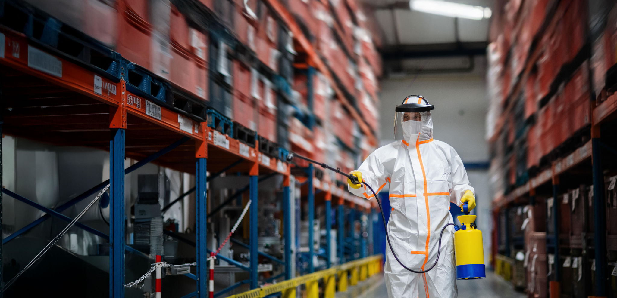 A person in protective gear sprays disinfectant along an aisle in a warehouse. They wear a white suit, face shield, gloves, and hold a yellow sprayer. Shelves lined with red containers are on both sides.