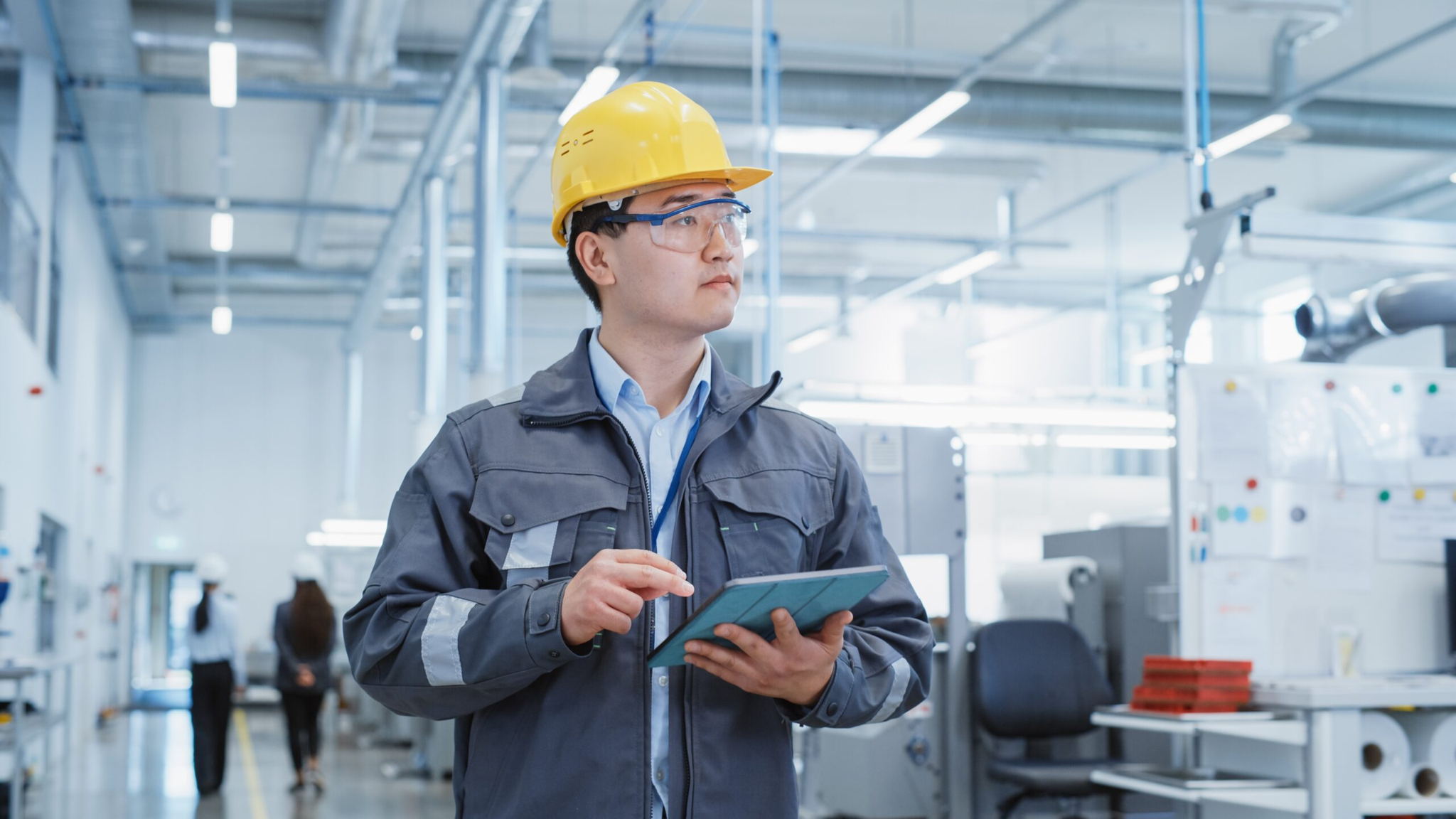 An engineer wearing a yellow hard hat and safety goggles uses a tablet in a modern industrial facility, with equipment and coworkers visible in the background.