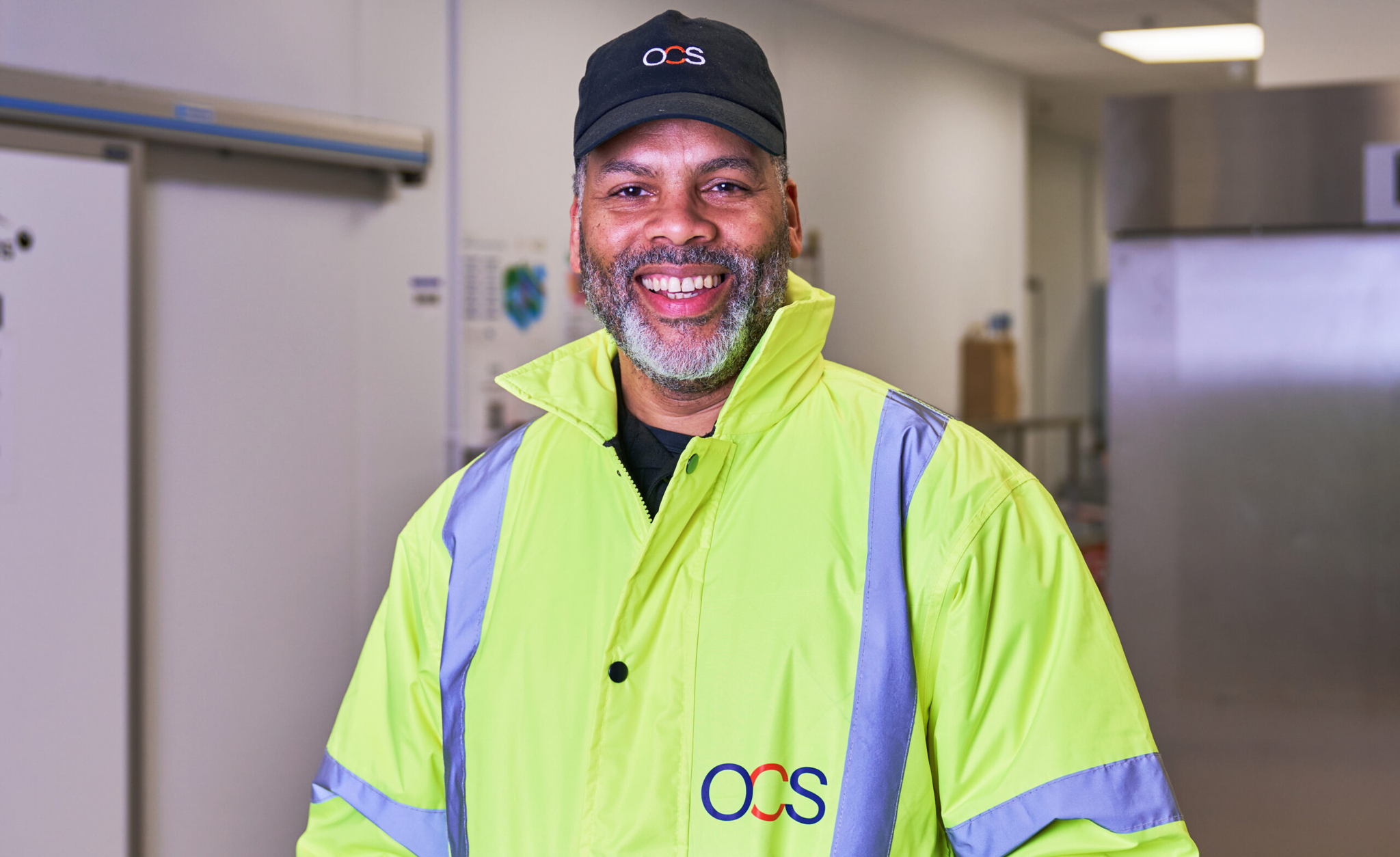 A smiling man wearing a high-visibility yellow jacket and a black cap, both with an OCS logo, stands indoors in a well-lit, modern setting.