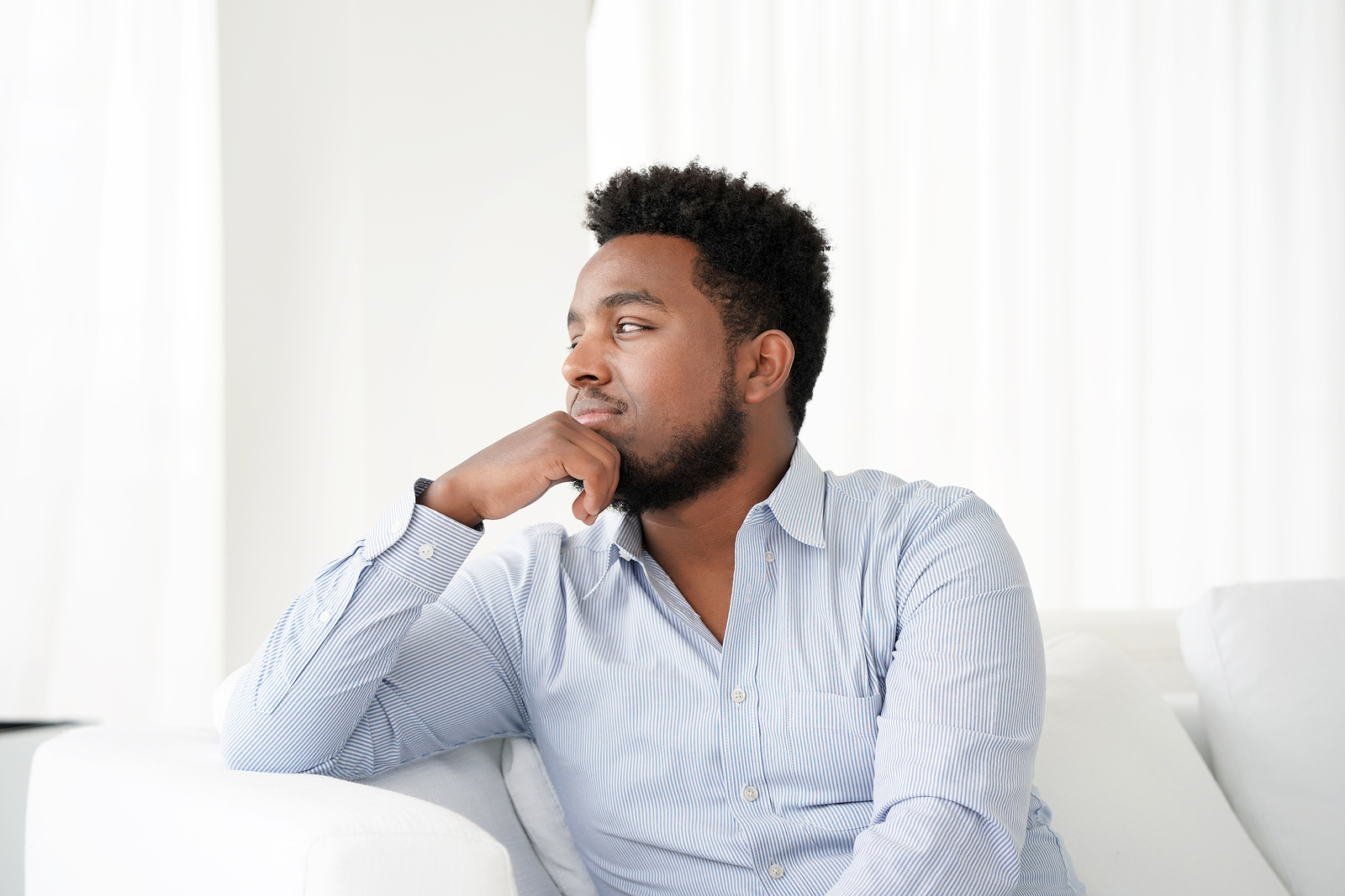 Photo of a  black man in a blue button up shirt looking pensive to the side on a white couch.