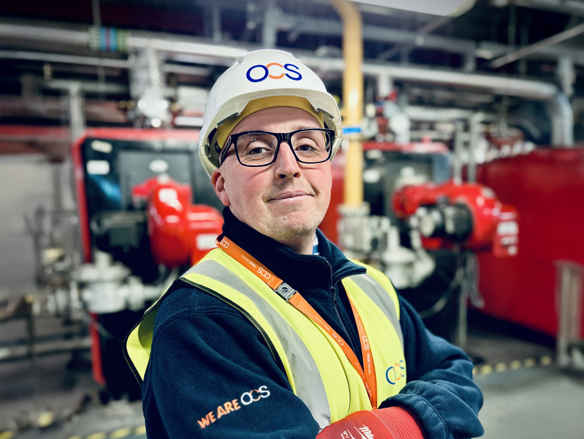 A confident worker in safety glasses, a white OCS helmet, and a yellow vest stands in an industrial setting, arms crossed, with machinery and pipes in the background.
