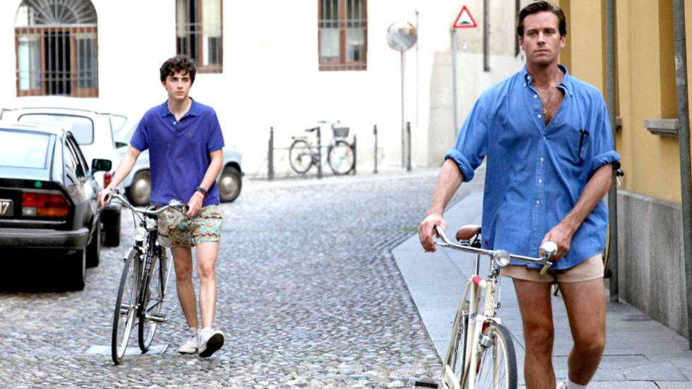 Elio staring at Oliver as they walk their bikes down a street. Both are wearing casual clothing and Oliver is walking ahead of him.
