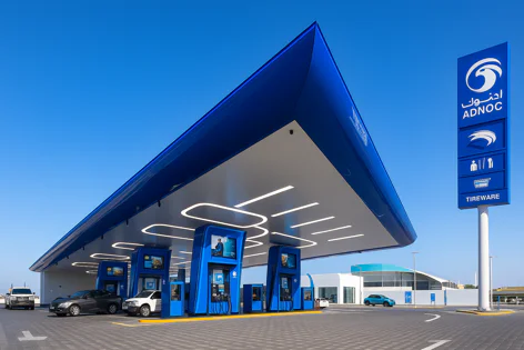 A modern ADNOC gas station featuring a large blue canopy, multiple fuel pumps, and cars refueling under a clear blue sky, with the prominent ADNOC sign.