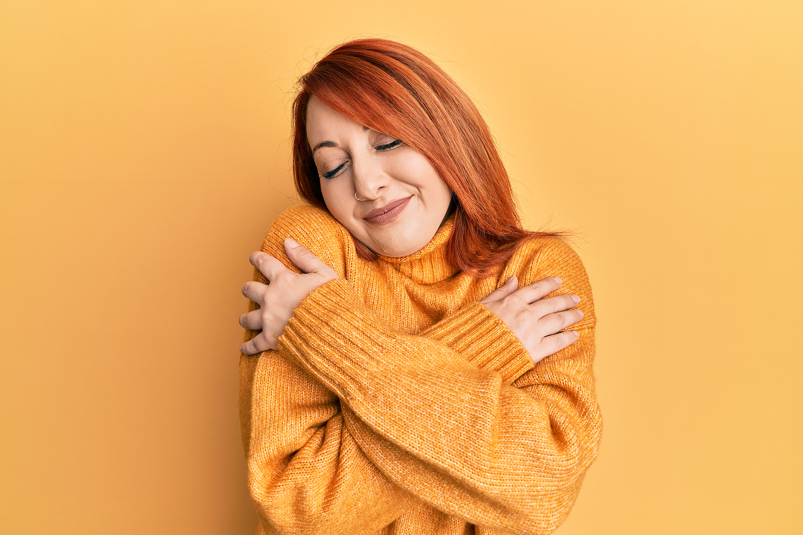 A woman with bright red hair and a warm sweater holds herself in a hug with her eyes closed.