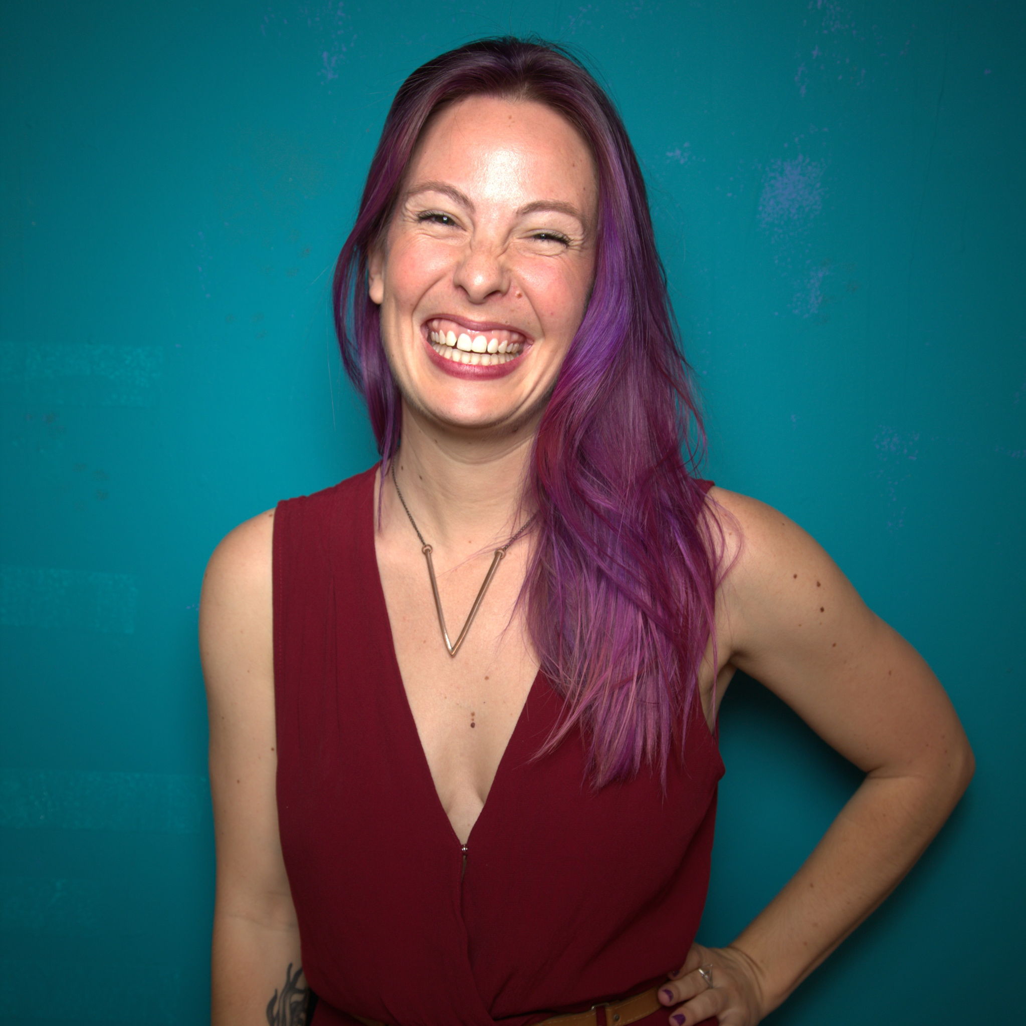 Photo of Amber Rollo wearing a red dress, purple hair and a big smile, against a teal background.