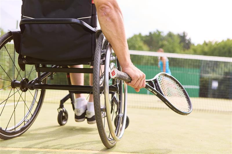 A person in a wheelchair holds a tennis racket on an outdoor tennis court, preparing to play. Another player stands in the background near the net.