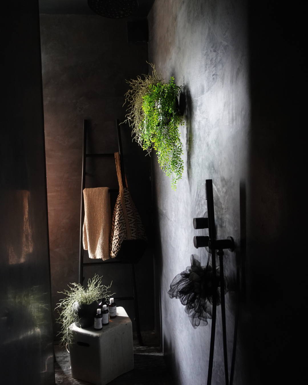 A walk in shower with black tadelakt walls, a lwooden ladder for towels and artificial plants.