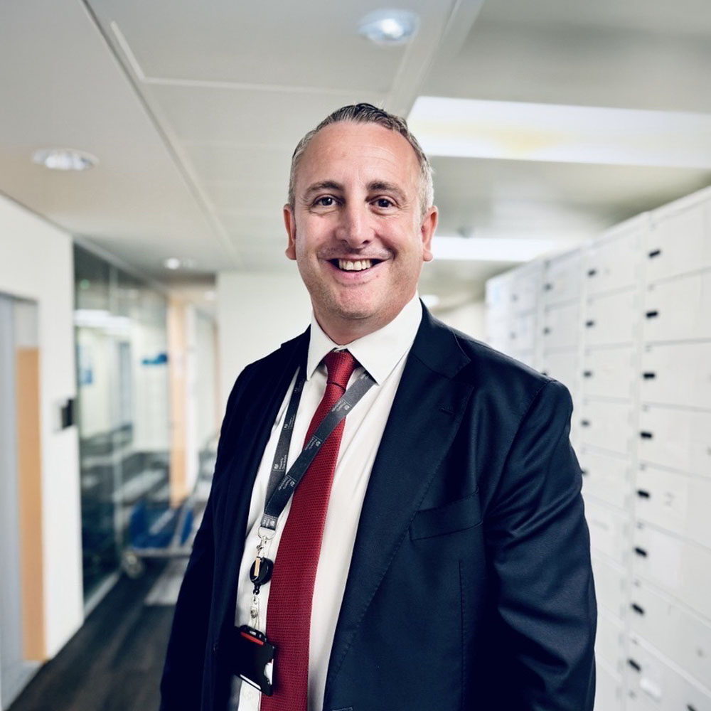 A man in a dark suit, white shirt, and red tie stands smiling in an office hallway with white cabinets and glass walls in the background. He wears a lanyard and has short, neatly styled hair.