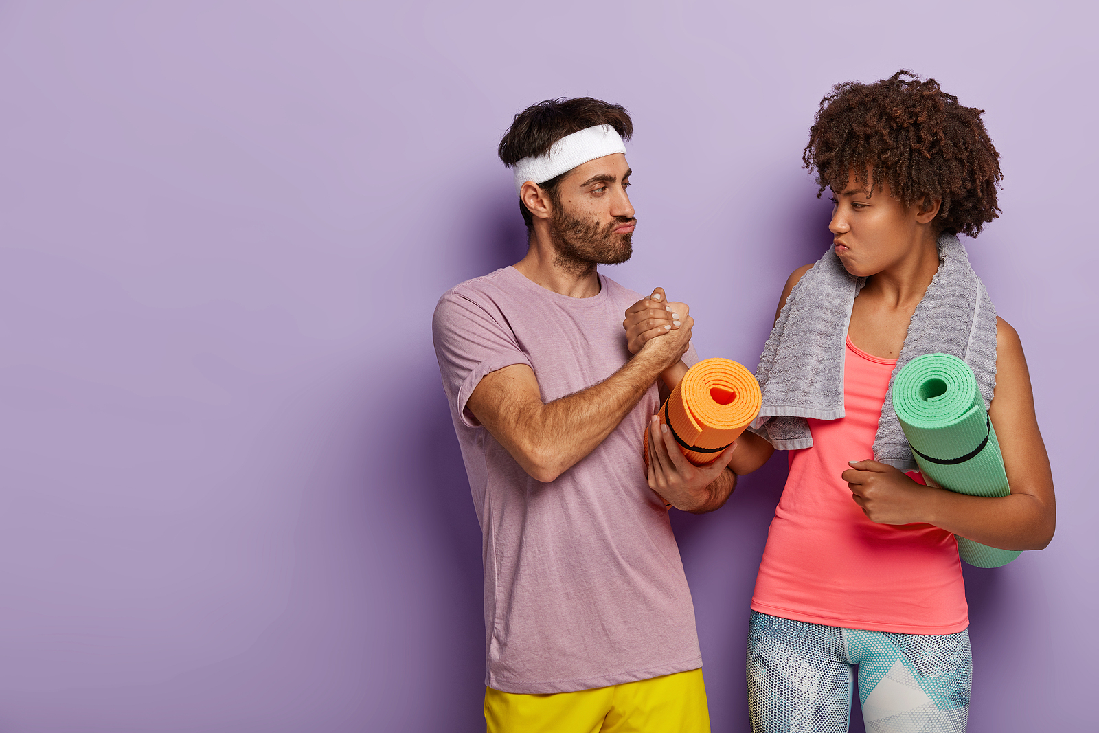 A confident man and woman wearing gym clothes lock arms and look at eachother with playful expressions.