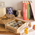 A box of spice jars on a kitchen table with cookbooks and spice mills behind them