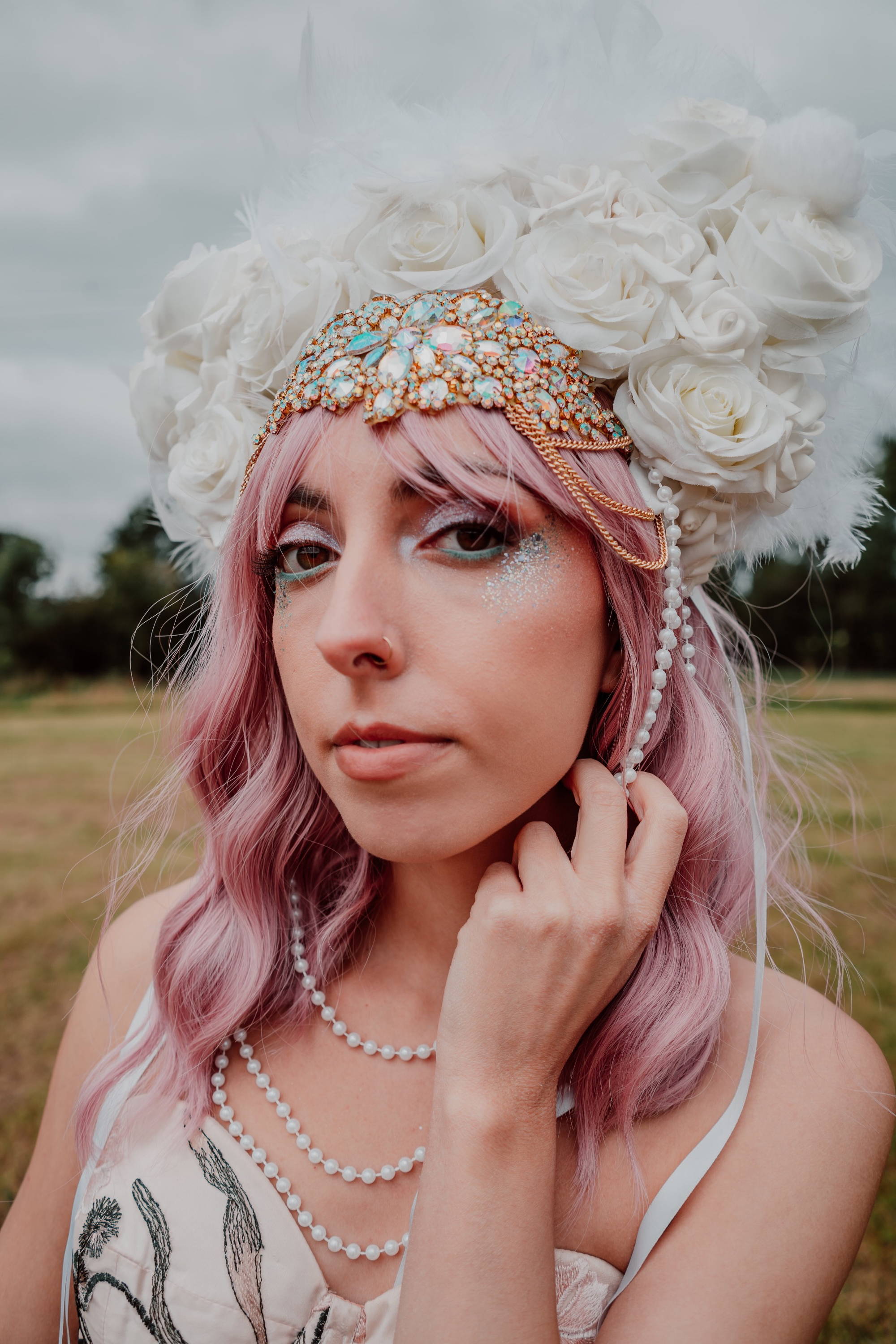 Image of a bride at a festival. She is wearing an embroidered modern bridal outfit, white head piece instead of a veil, and has biodegradable blue, pink, and iridescent glitter on her face. 