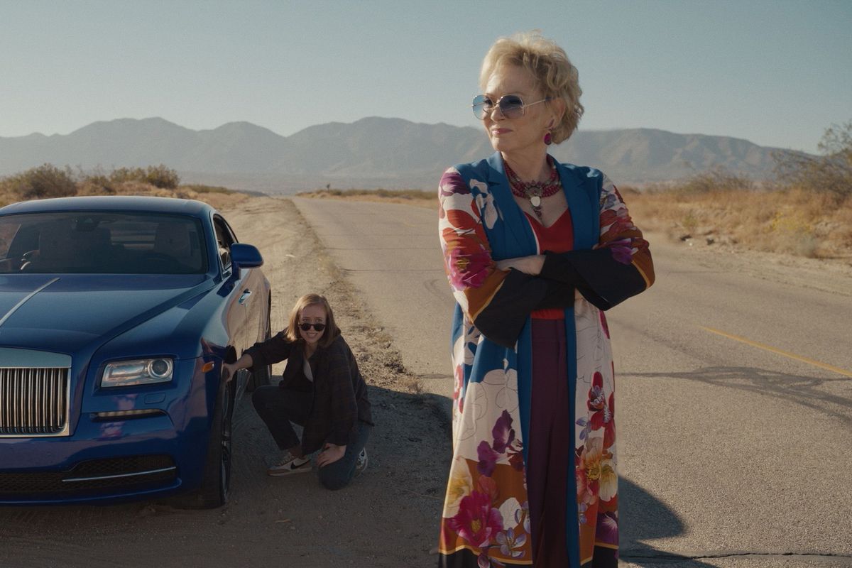 Ava is kneeling near the blue car and Deborah, with her arms crossed, looks off to the distance with a smile.they are on the side of the road.