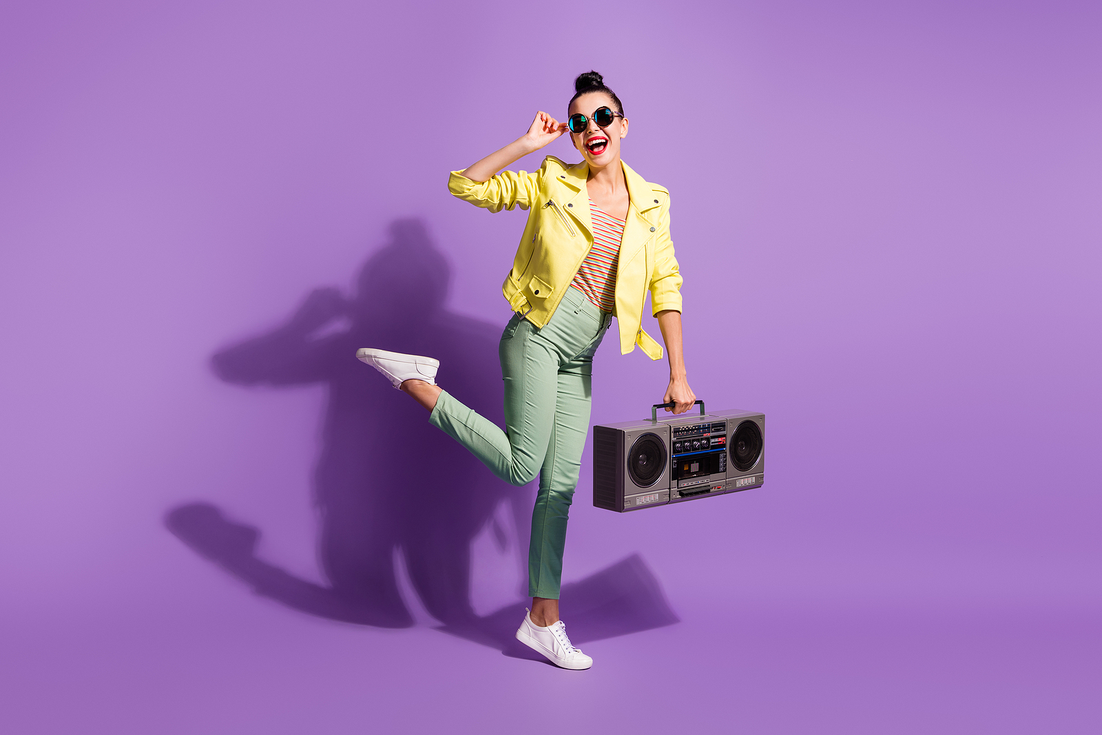 A cheerful woman with sunglasses poses with a boombox, smiling and with a leg in the air.