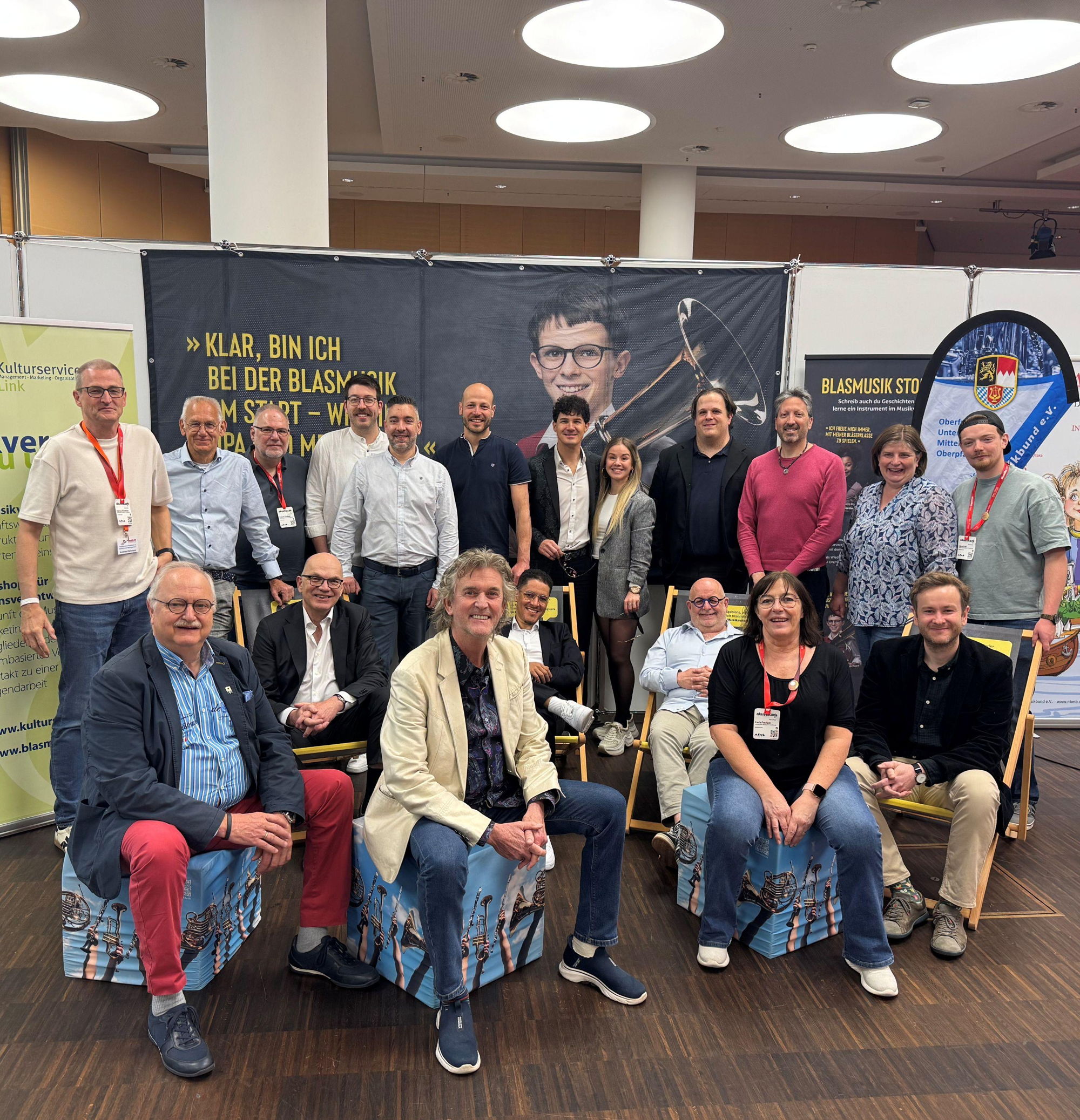 Viele Männer und einige Frauen stehen und sitzen auf einer Messe beieinander und lächeln in die Kamera. 