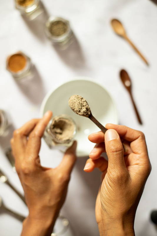 Ingredients for chai masala being scooped into a white bowl with a small wooden spoon