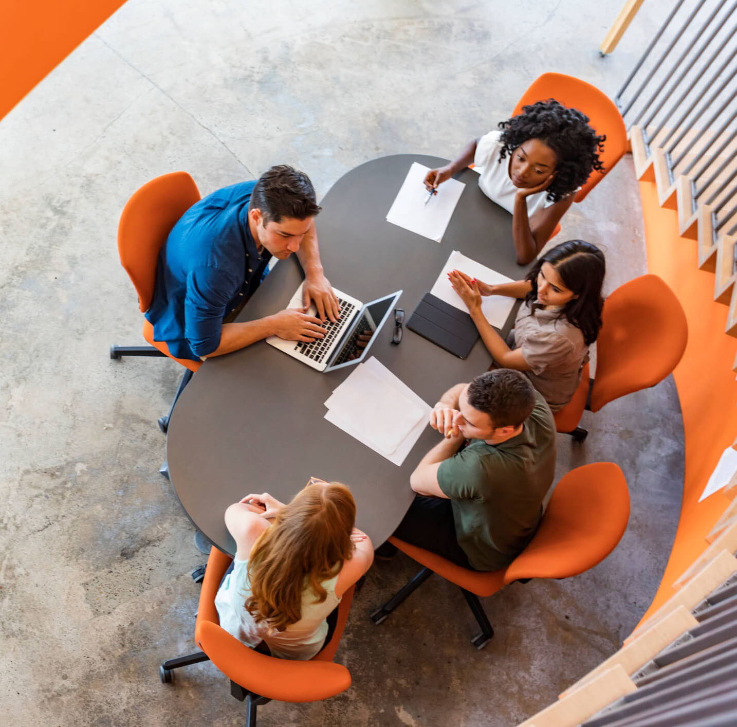 Aerial view of five people engaged in a meeting around an oval table. One person is using a laptop, while others have notebooks and papers. The chairs are orange, contrasting with the gray table. The setting is modern and collaborative.
