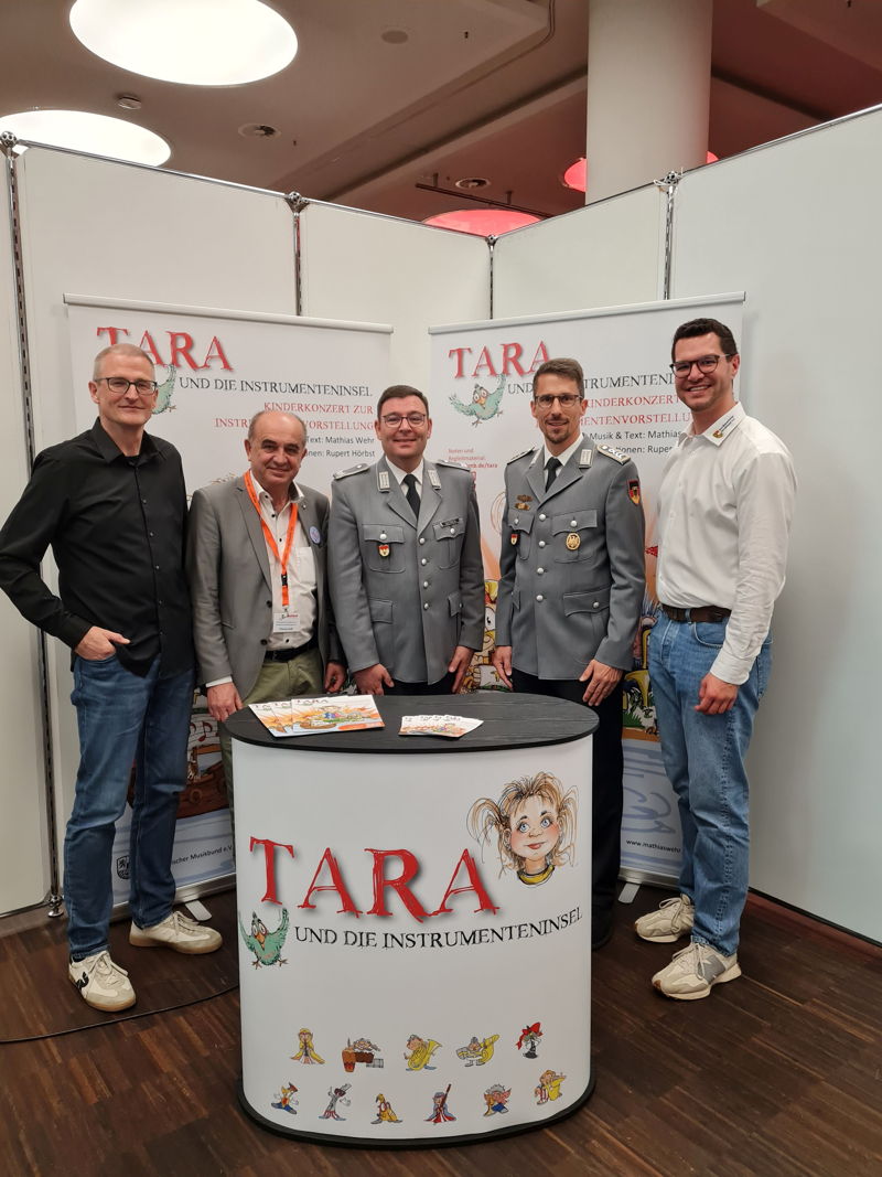 Fünf Männer, zwei von ihnen in Uniform, stehen beieinander und lächeln in die Kamera. Vor ihnen steht eine Messetheke und hinter ihnen Roll-ups, auf denen "Tara und die Instrumenteninsel" zu lesen ist. 