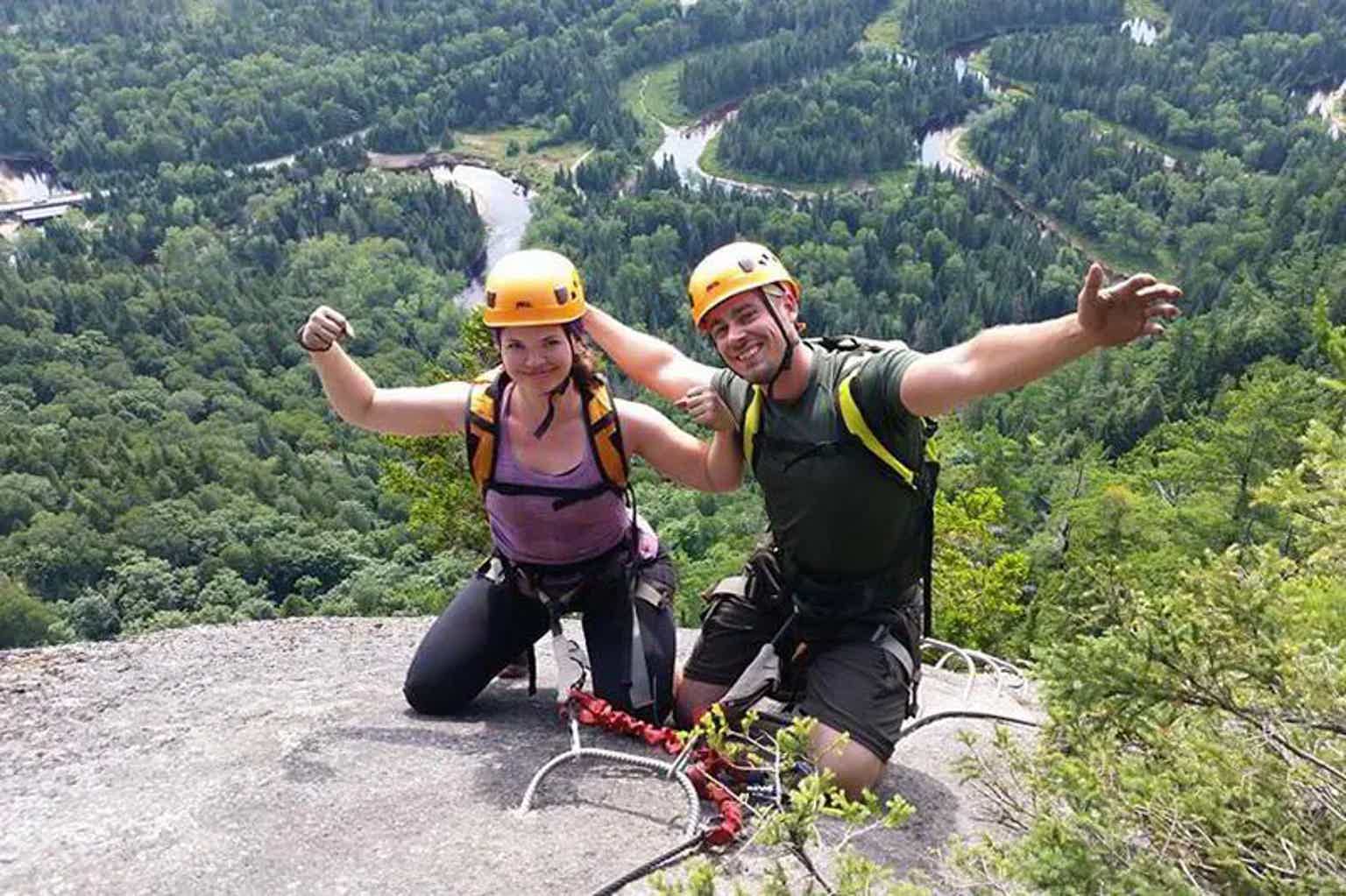 Two climbers on a rocky overlook wearing helmets, harnesses, posing with arms raised, lush forest and winding road below.
