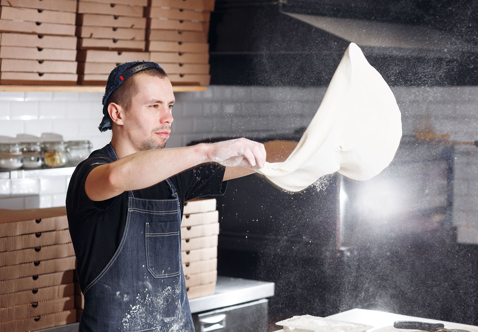 A man with a backward baseball cap working at a pizzeria tossing dough in the air.