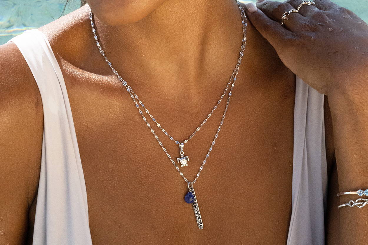 A women's neck with two layered neckalces; one with a turtle charm and one with a coordinates charm.