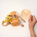 A hand holding a wooden disc that says "Hello World" with a crochet giraffe toy, a blanket, brush, and a pacifier below. 