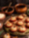 Round Indian sweets topped with whole almonds in red paper liners on a golden tray, surrounded by earthen bowls and honey in the background.