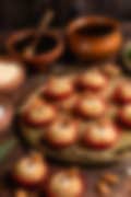 Round Indian sweets topped with whole almonds in red paper liners on a golden tray, surrounded by earthen bowls and honey in the background.