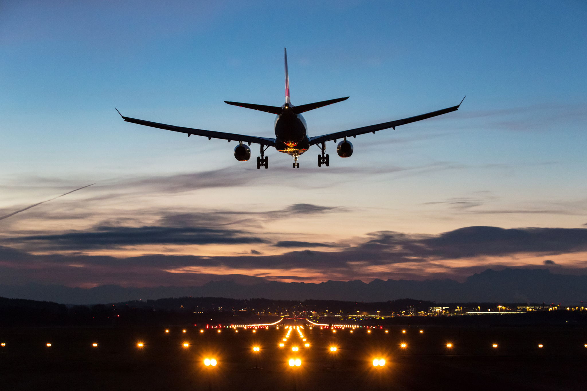 A commercial airplane prepares to land on a runway at dusk, with runway lights illuminated and a colorful sky with clouds in the background.