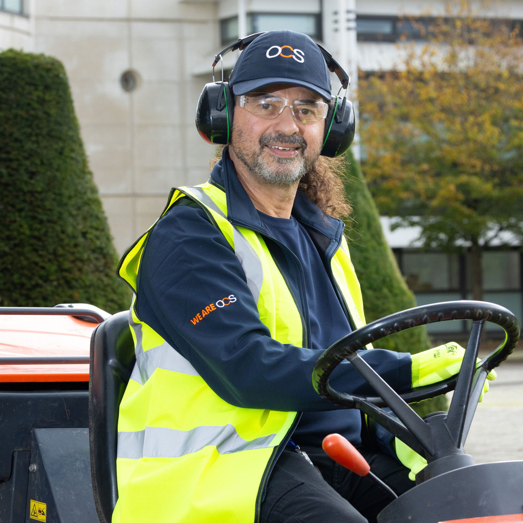 A man wearing a high-visibility vest, safety glasses, ear protection, and an OCS cap sits smiling at the wheel of an outdoor vehicle, with trees and a building in the background.