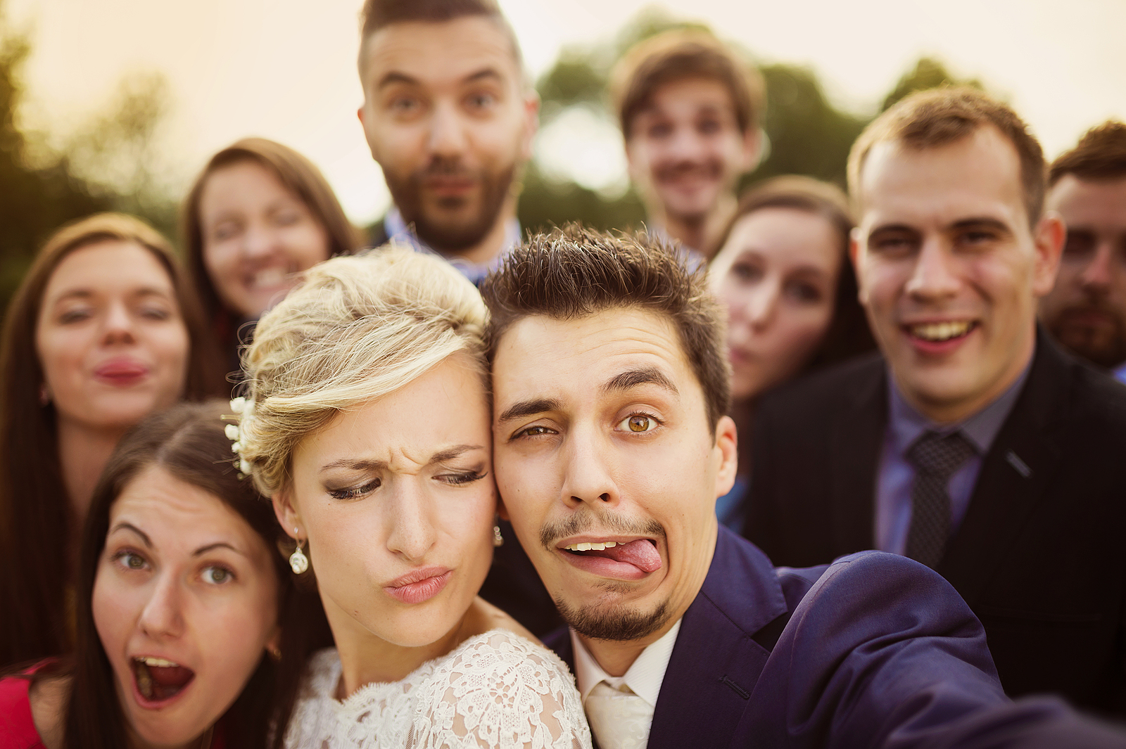 A bride and groom making silly faces in the camera with a group of friends smiling in the background