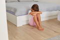Child sitting on the floor beside a bed with head down and arms wrapped around their knee.
