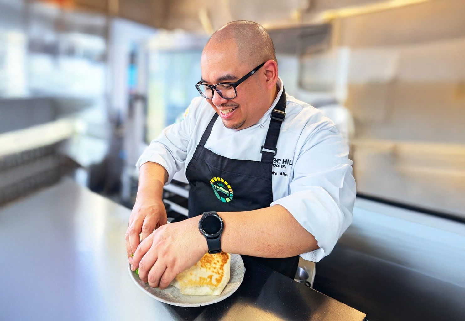 A smiling chef wearing glasses and a black apron prepares a dish on a countertop in a bright, modern kitchen.
