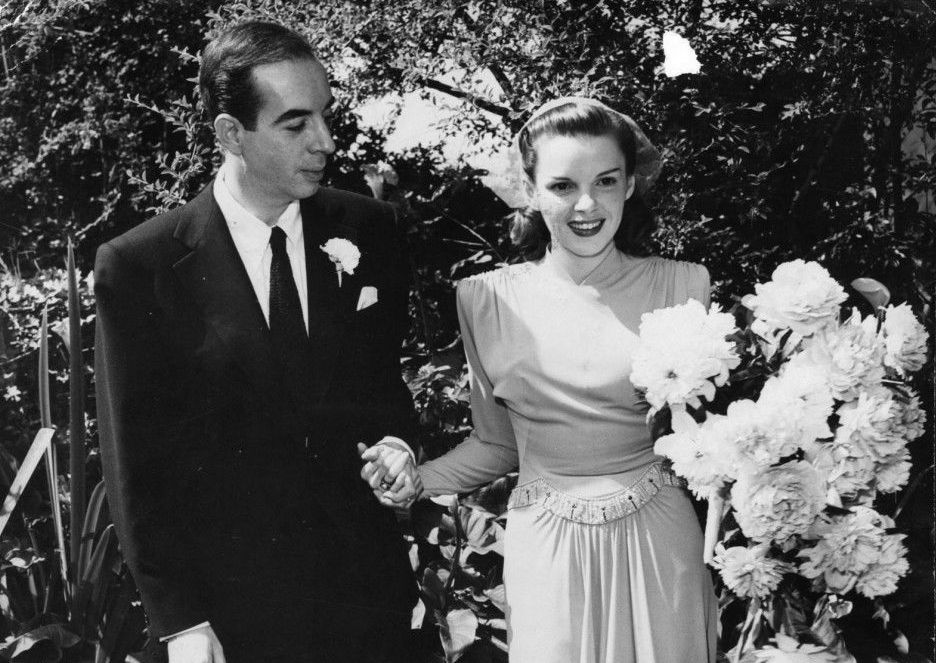 Vincent Mineli in a suit holding the hand of a smiling Judy Garland in a white dress with a large bouquet on their wedding day.