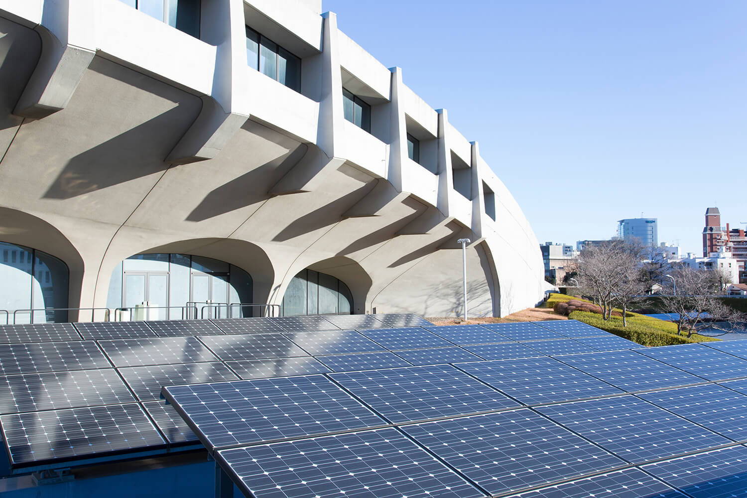 Rows of solar panels are installed outside a modern, curved concrete building under a clear blue sky, with city buildings visible in the background.