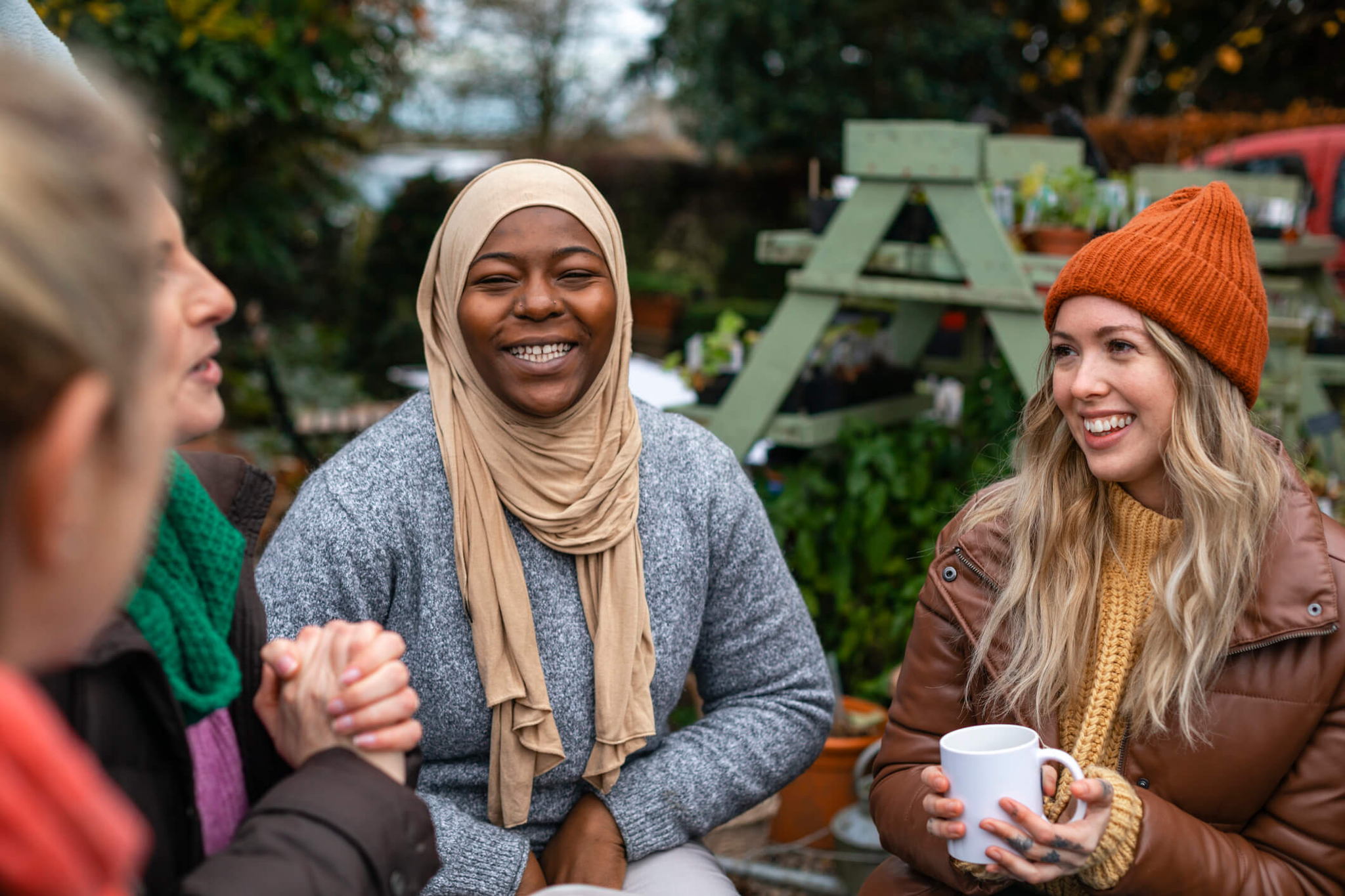 A group of people are sitting outside, smiling and enjoying hot drinks. The setting includes plants and wooden structures in the background. The atmosphere appears relaxed and friendly, with everyone dressed warmly for cold weather.