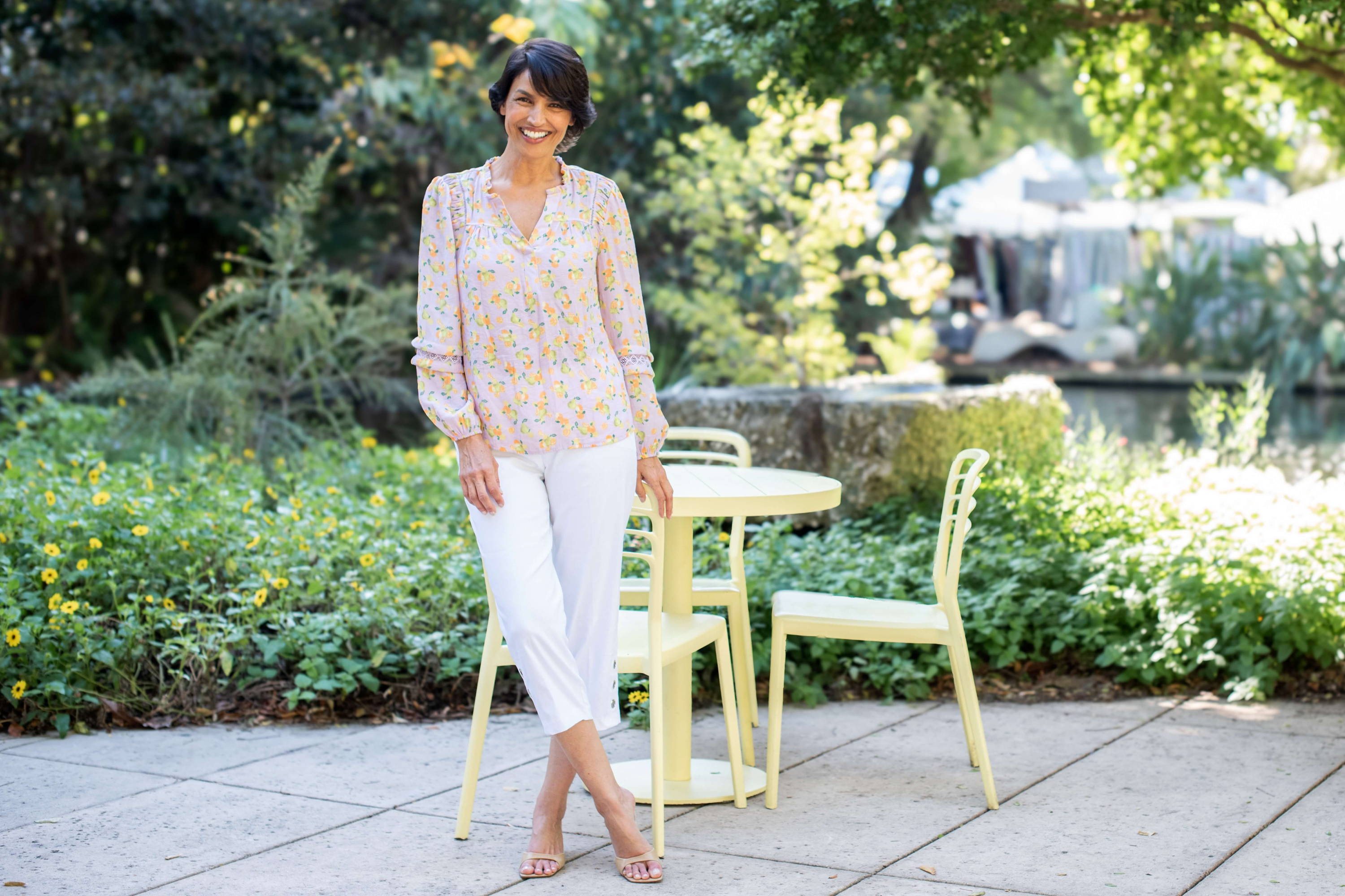 A woman wearing a floral blouse and white capri pants stands in front of a bistro table on a garden patio