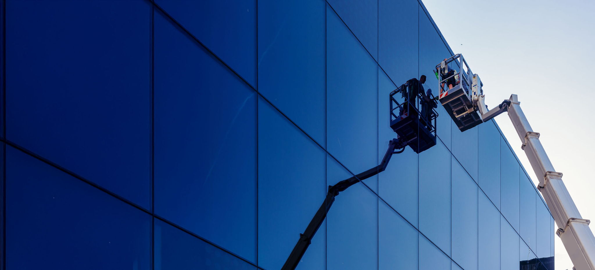 Two workers in lift buckets clean or inspect the windows of a modern glass building, reflected in the blue-tinted panels under a clear sky.