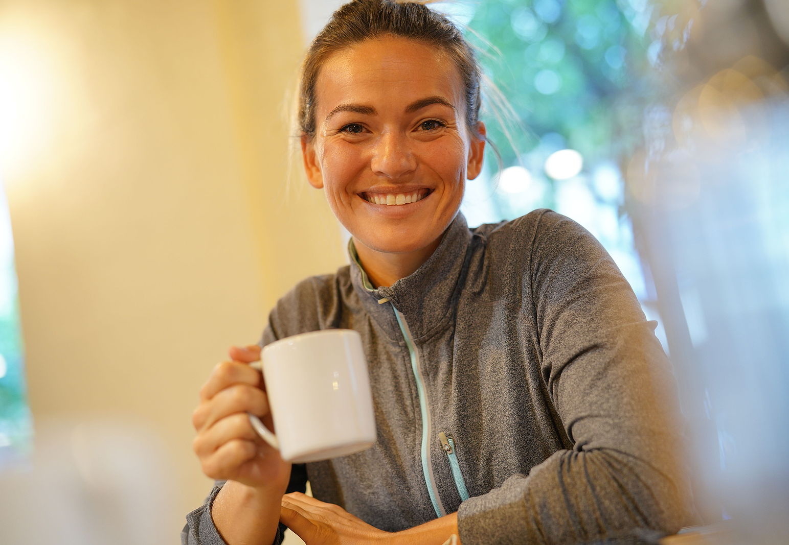 An older blond haired woman with her hair up holds her coffee cup and smiles big for the camera.