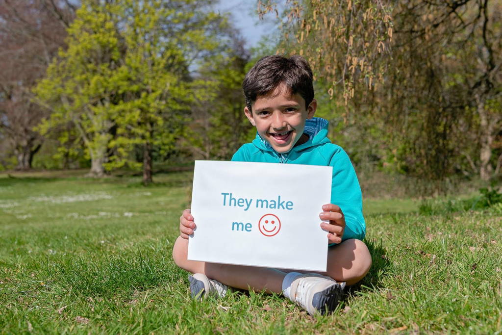 Image of smiling boy sitting outside holding a sign that says 'They make me happy'. The boy is wearing Ducky Zebra clothes.