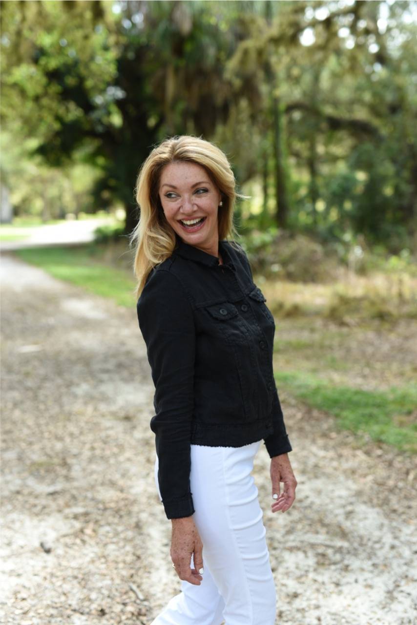 A woman wears a black jacket with white denim pants in a sunny yard.