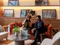 Zwei Personen sitzen auf einem orangefarbenen Sofa in einer stilvollen Hotellobby mit Marmor-Couchtisch.