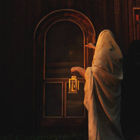 Jesus standing at a door and knocking. Hisi surroundings are dim and He carries a lamp.